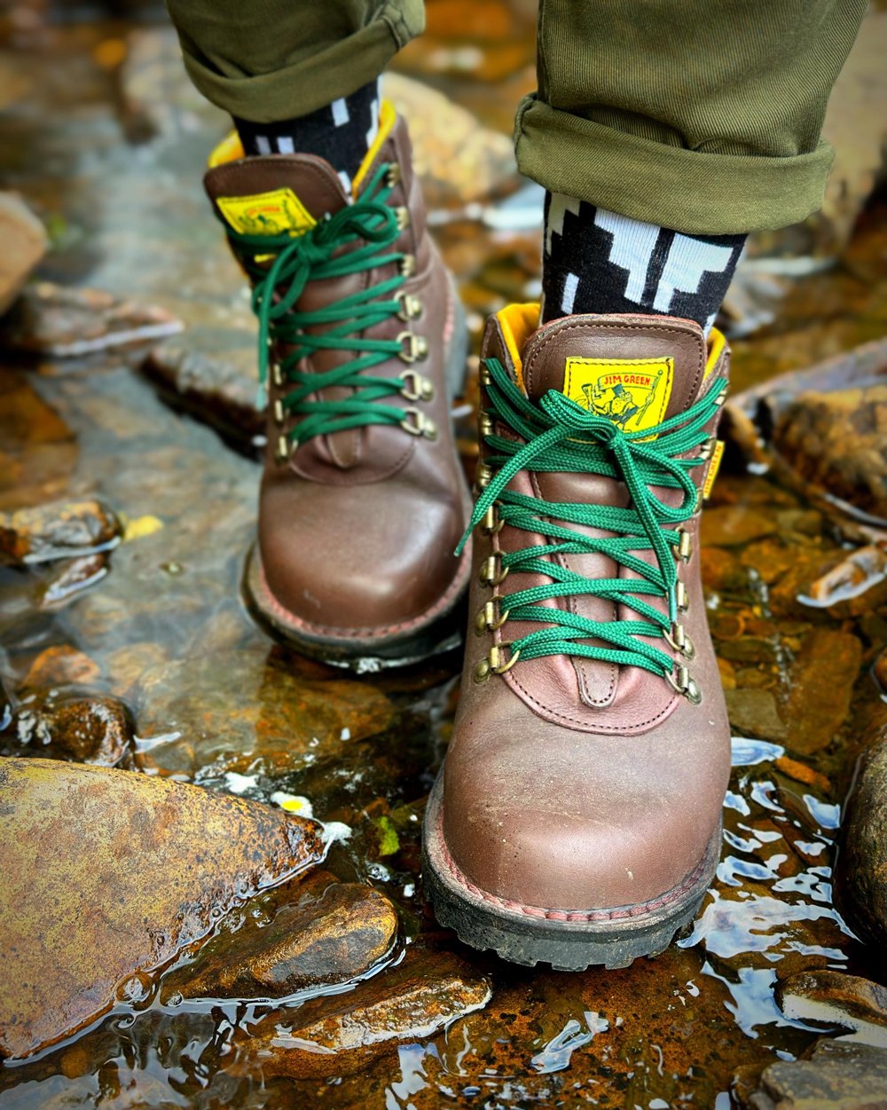 Close up of brown Jim Green boots with green laces standing in a shallow rocky stream