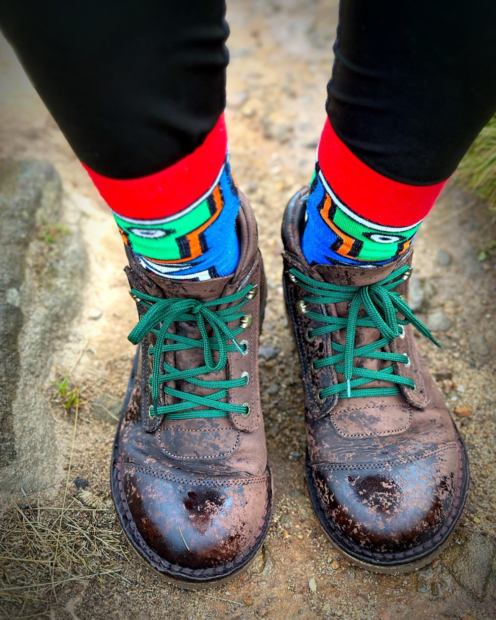 Close-up of worn brown leather boots with bright green laces and colorful patterned socks on a dirt path